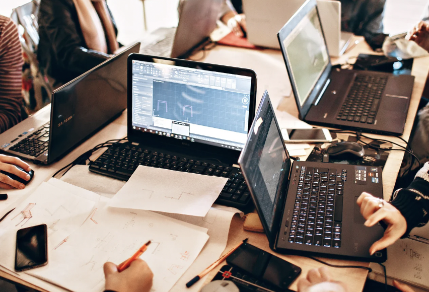 a conference table with several laptops, individuals around table, but mostly not depicted except for a few hands writing notes.