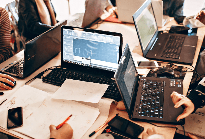 a conference table with several laptops, individuals around table, but mostly not depicted except for a few hands writing notes.